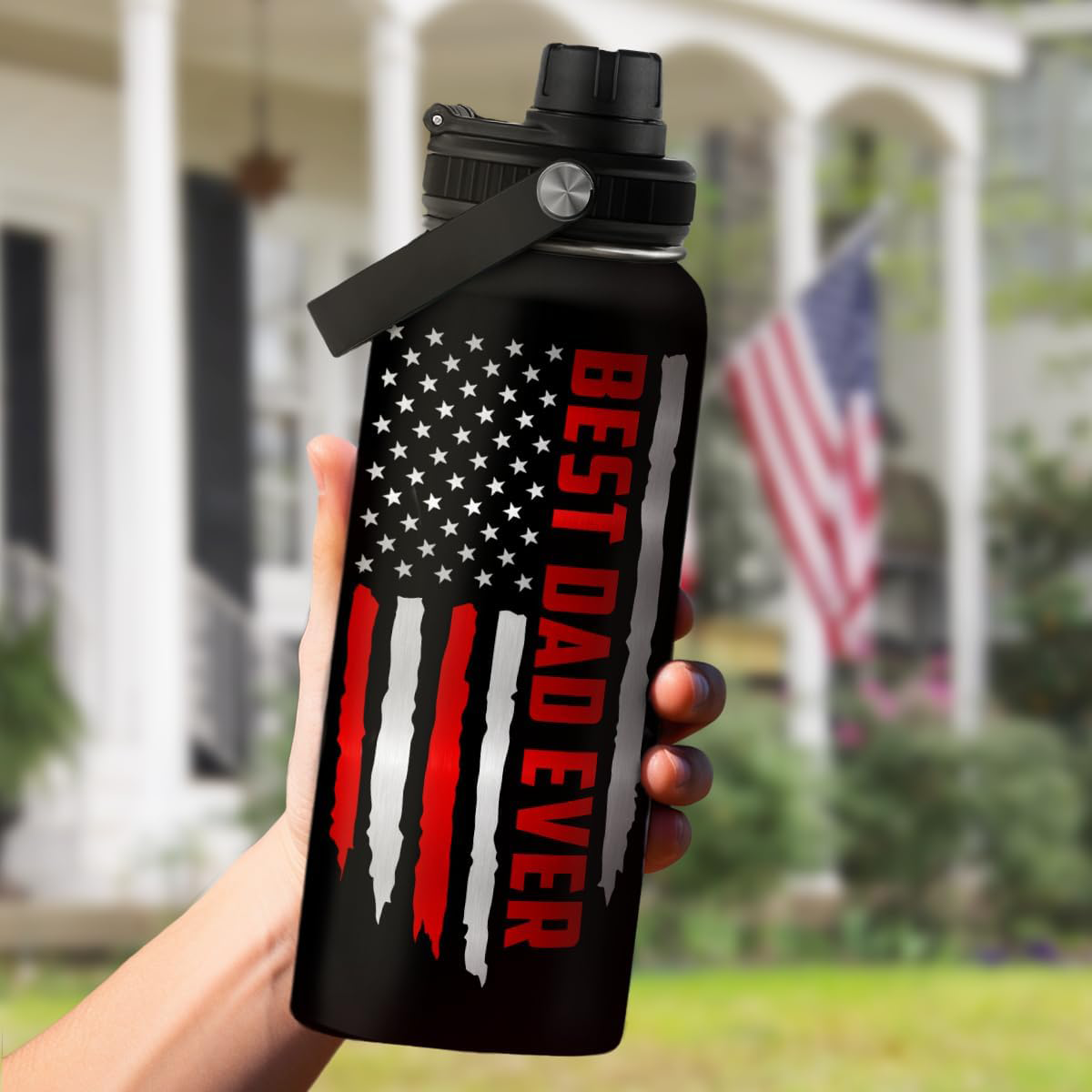 Black water bottle with American flag design and 'Best Dad Ever' text, held in front of a house with an American flag.