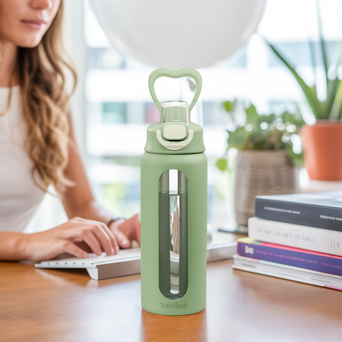 Green water bottle with a clear window on a desk with a person using a laptop and books in the background.