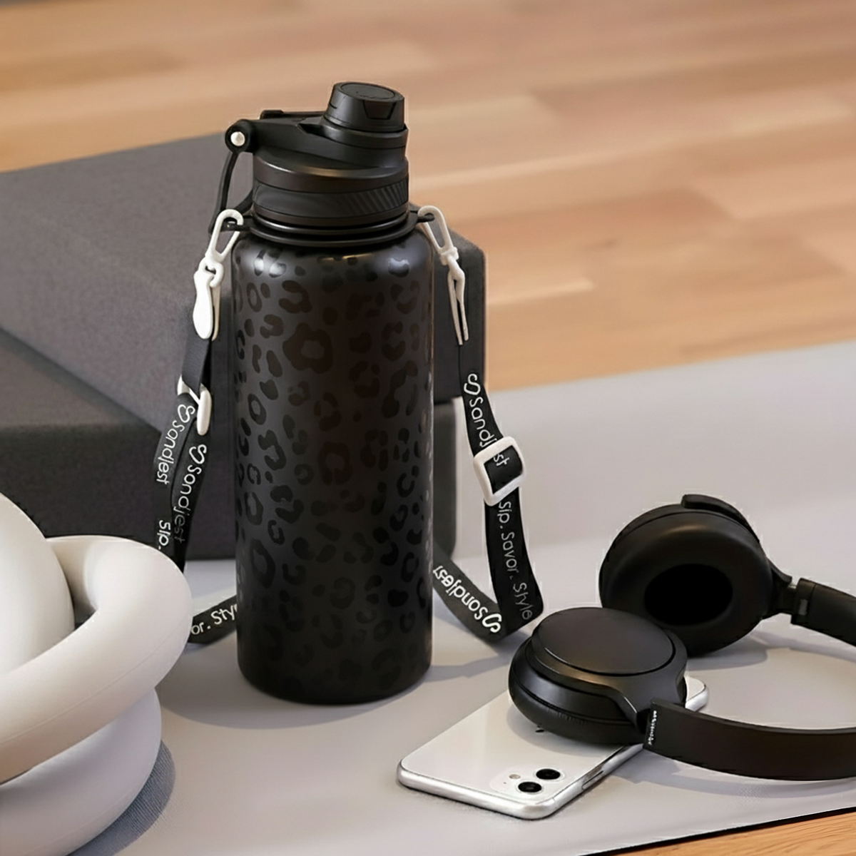 Black water bottle with leopard print pattern on a wooden surface, surrounded by a phone and headphones.