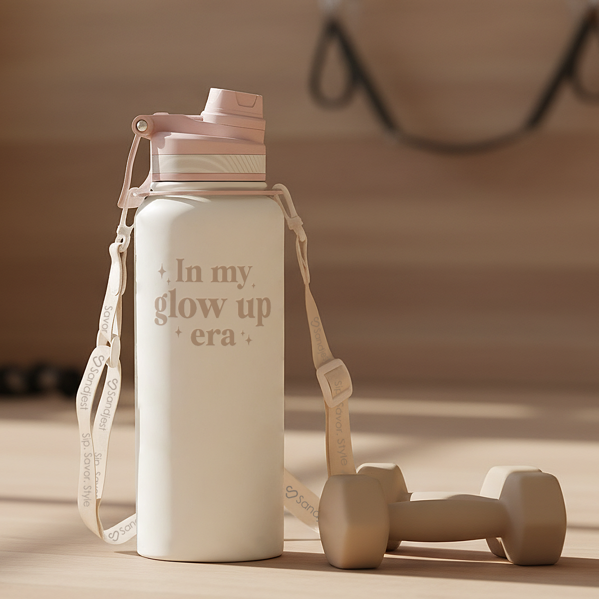 White water bottle with 'In my glow up era' text, pink cap, and white strap on a wooden surface with dumbbells.