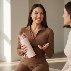 Woman in brown outfit holding a beige water bottle, smiling and interacting with another person.