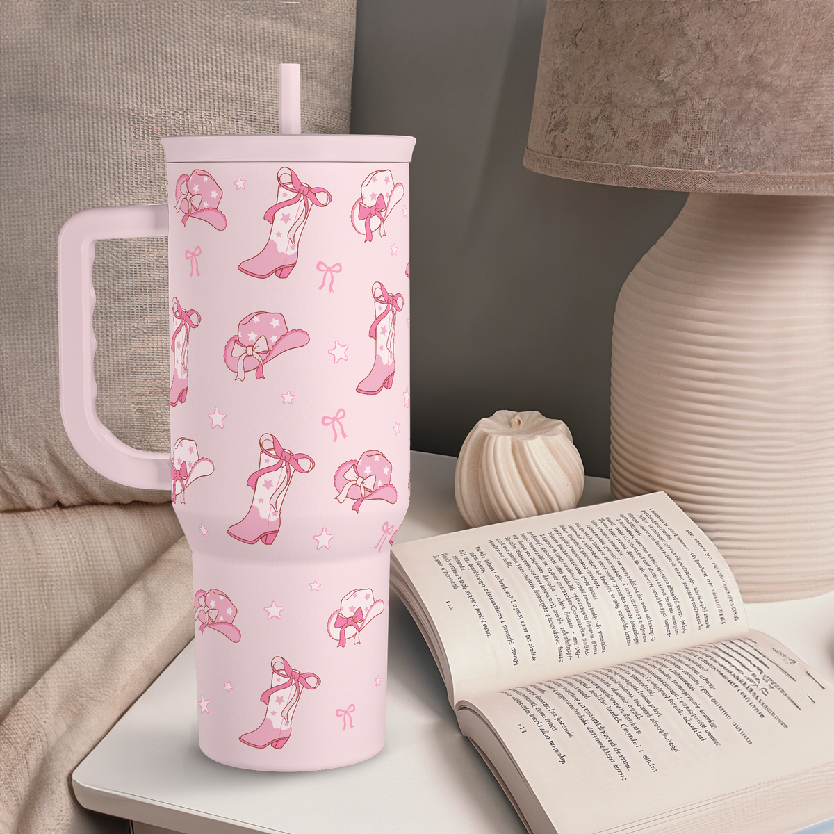 Pink tumbler with shoe design on a surface next to an open book and lamp.