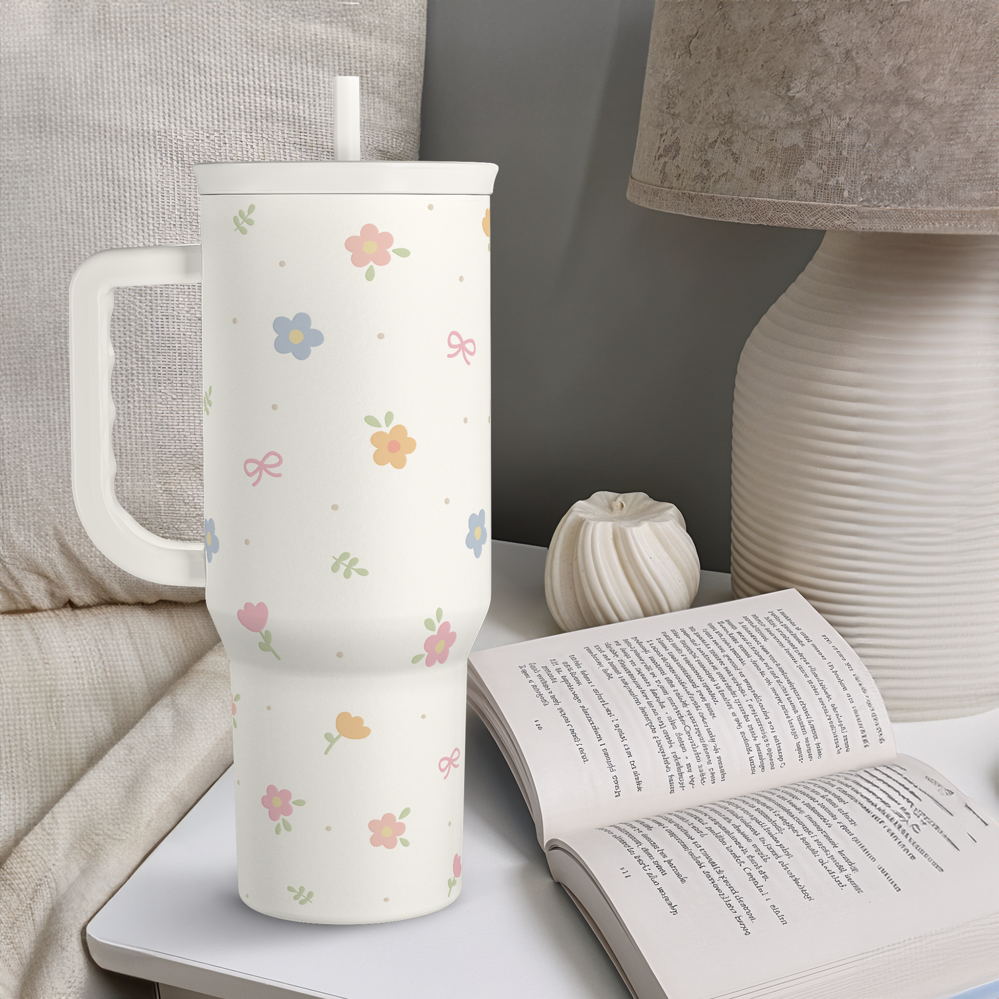 White mug with floral pattern on a table next to an open book and decorative lamp.